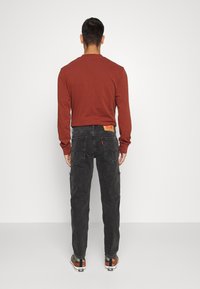 Camisa de manga larga color óxido combinada con pantalones cargo negros que tienen bolsillos laterales y un parche de la marca. Zapatos con un diseño de rayas multicolores.