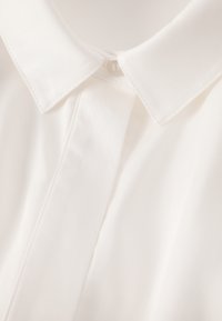 Biała koszula z kołnierzykiem, wykonana z gładkiej tkaniny, z ukrytym plisowaniem na guziki oraz starannym przeszyciem wzdłuż kołnierza i przedniego panelu.