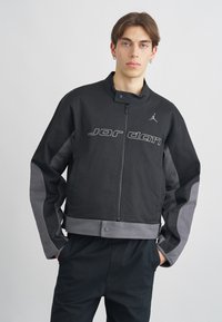 Casaco bomber Jordan em preto e cinza, com fecho frontal, gola alta e detalhe do logótipo, apresentando um design cropped e acabamentos em tecido texturizado.