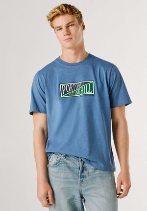Blaues Baumwoll-T-Shirt mit einem Logodesign, das "Portobello Notting Hill" in Grün und Weiß zeigt, kombiniert mit hellen Denim-Jeans.
