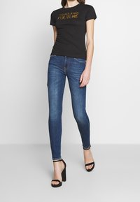 Žena v černém tričku Versace Jeans Couture, modrých skinny džínách a černých sandálech na vysokém podpatku na bílém pozadí.