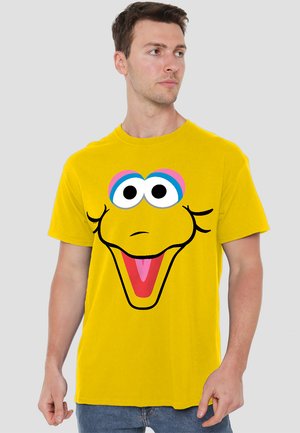 Gelbes T-Shirt mit dem Gesicht eines Cartooncharakters, der große Augen, ein breites Lächeln und schwarze Konturen hat. Hergestellt aus Baumwolle, kurzärmliges Design.