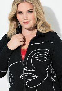 Czarna kurtka zapinana na zamek z białym rysunkiem twarzy. Kurtka ma fakturowaną tkaninę i jest noszona na czerwonym topie.