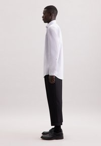 Camicia bianca a maniche lunghe, con polsini abbottonati, indossata con pantaloni neri su misura e scarpe nere con lacci. Design semplice e pulito con una vestibilità classica.