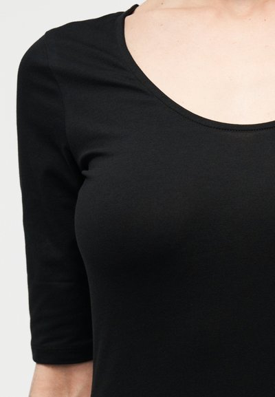 Κοντινό πλάνο ενός ατόμου που φοράει εφαρμοστή μαύρη μπλούζα με λαιμόκοψη σχήματος scoop και μανίκια τριών τετάρτων, σε ανοιχτό φόντο.