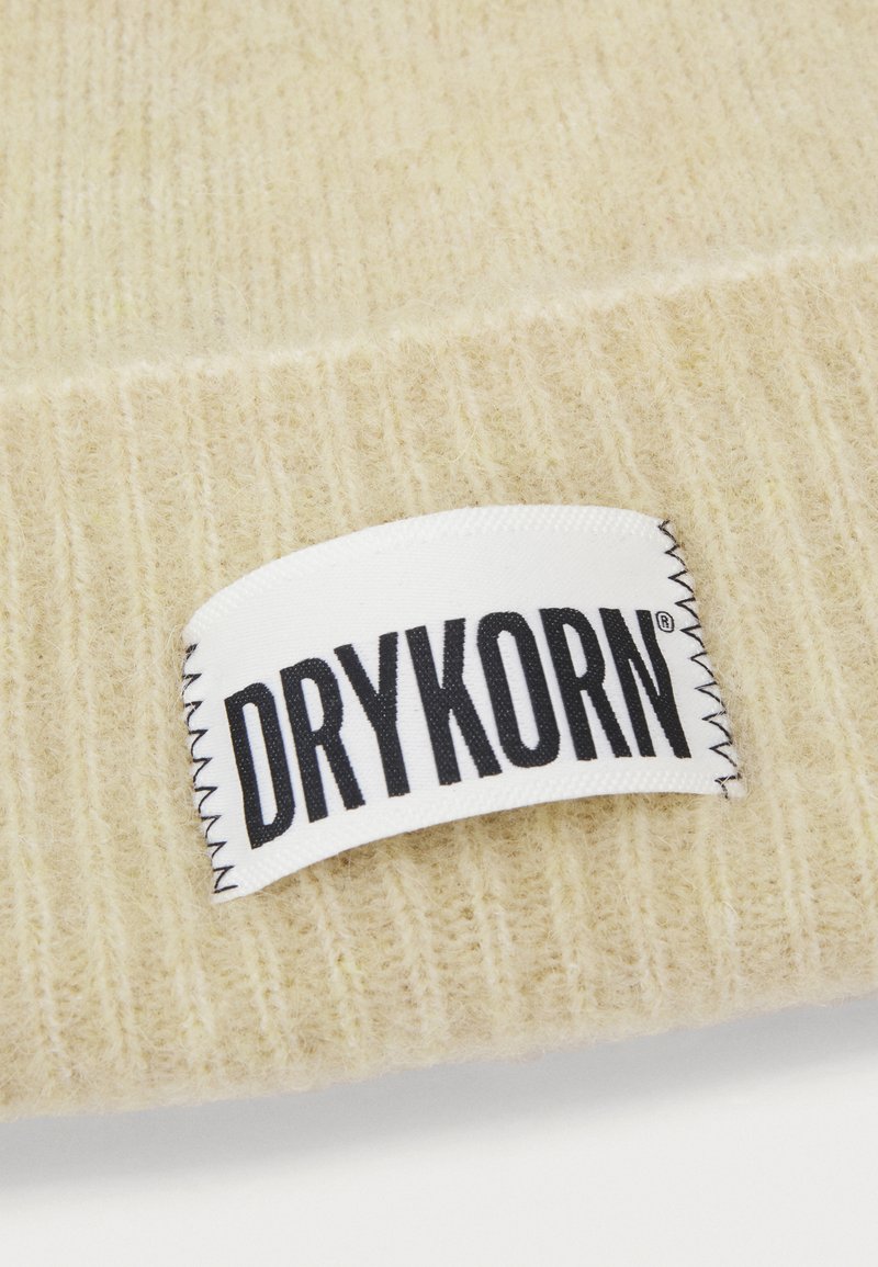 Beige gestrickte Mütze mit strukturiertem Muster; hat ein weißes Etikett mit "DRYKORN" in Schwarz, das vorne mit sichtbarem Stich angenäht ist.