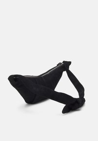 Sac en suède noir avec une forme triangulaire et une sangle nouée. Il dispose d'une fermeture éclair et d'une texture lisse avec des détails de surpiqure subtils.