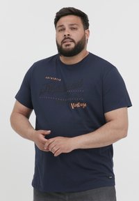 Mørkeblå bomulls T-skjorte med korte ermer, med trykt tekst i svart og oransje, samt en liten logodetalj nær kanten.