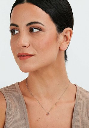 Goldene Halskette mit einem kleinen, runden, roten Edelstein-Anhänger. Das Model trägt ein beiges, geripptes Oberteil, das den Schmuck deutlich vor einem neutralen Hintergrund zur Geltung bringt.