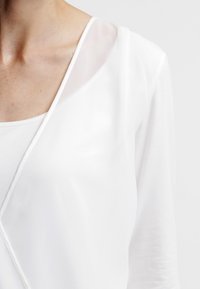 Blouse blanche avec un décolleté en V, superposition transparente et manches longues, fabriquée à partir d'un tissu léger au toucher lisse.