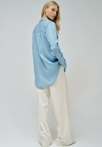 Camisa de mezclilla azul claro con mangas largas y cuello desmontable, combinada con pantalones blancos holgados, sobre un fondo neutro.