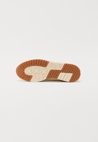 Såle av en sko med et teksturert design, lys gummitraksjonsmønster og en kremfarget base med riller og merkevaredetaljer.