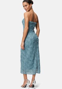 Robe bleue sans bretelles avec une texture en dentelle florale, longueur genou, fente sur le côté, portée avec des sandales à talons hauts argentées. Présente une silhouette cintrée.