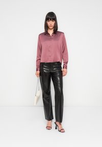 Langsleeve, button-up shirt in mauve satijn, gecombineerd met hooggesneden, glanzende zwarte faux leren broek. Model houdt een crèmekleurige handtas vast.