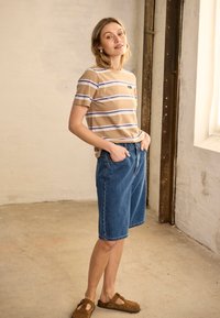 Femme debout à l'intérieur portant un t-shirt beige à rayures, un short en jean longueur genou et des chaussures marron sans lacets, les mains dans les poches.