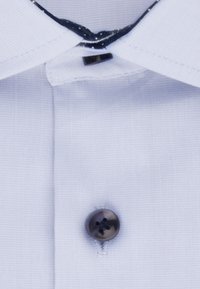 Camicia bianca con una trama liscia, caratterizzata da un bottone scuro e un colletto a contrasto. Include dettagli di cucitura attorno alla pattina dei bottoni.