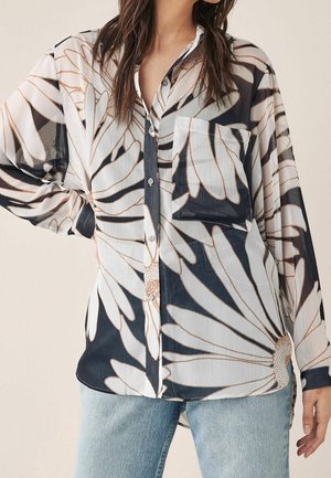 Femme portant une chemise transparente boutonnée avec de grandes imprimés marguerites blanches et un jean bleu clair, debout avec une main sur la hanche.