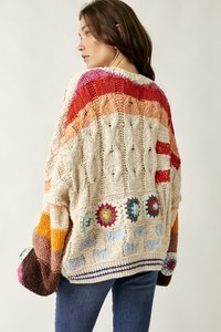 Γυναίκα με μπλε τζιν που φοράει ένα φαρδύ, πολύχρωμο πλεκτό πουλόβερ με υφασμένους σχεδιασμούς και κυκλικά μοτίβα στην πλάτη.