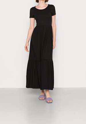 Woman wearing a black short-sleeve, ankle-length dress and purple woven open-toe slide sandals, standing on gray floor against white wall.