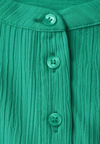 Grön, texturerad tyg med vertikala plisseringar, prydd med tre runda plastknappar märkta "CECIL" framtill.