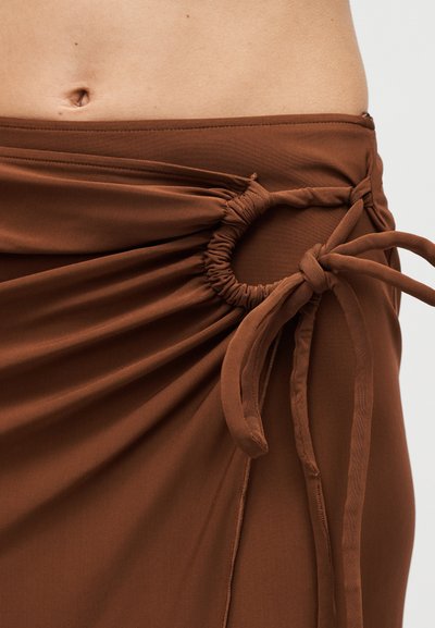 Καφέ υφασμάτινη φούστα με λεπτομέρεια πτυχώσεων στο πλάι δεμένη με φιόγκο, φορεμένη στη μέση και αποκαλύπτει μερικώς την κοιλιά και τον αφαλό.