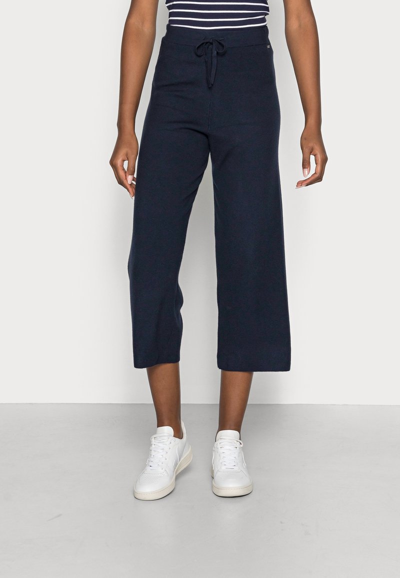 Femme portant un pantalon large cropped bleu marine avec taille à cordon et des baskets blanches, se tenant sur un sol gris.