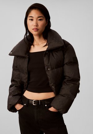 Jonge vrouw met een korte zwarte donzen jas, een zwart cropped topje en zwarte broek met handen in de zakken, staand tegen een effen achtergrond.