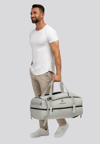 Homme en t-shirt blanc et pantalon beige tenant un sac de sport gris clair avec des zippers noirs et le logo "pactastic."