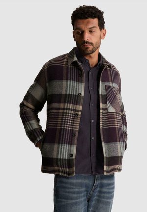 Homme portant une veste à carreaux violette et grise sur une chemise sombre boutonnée et un jean bleu, les mains dans les poches de la veste, regardant vers le bas.