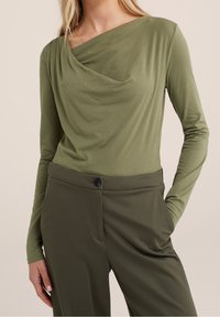 Long-sleeve, draped top in olive green fabric, featuring a cowl neckline and smooth texture. Paired with tailored olive-green pants.