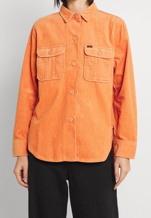 Person wearing a buttoned orange corduroy shirt with two chest pockets over black pants, standing against a plain light background.