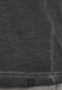 Tricou din bumbac gri cu un aspect texturat și purtat. Are o etichetă din material negru în colțul din stânga jos. Mânici simple, scurte.