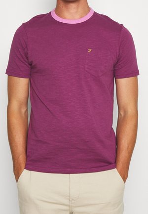 Homme portant un t-shirt violet à manches courtes avec un col rose et une petite poche poitrine, les mains dans les poches d'un pantalon beige.