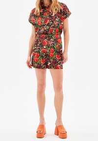 Mono con estampado tropical de manga corta, que presenta frutas rojas y follaje verde sobre un fondo negro. Combinado con sandalias de plataforma naranjas.
