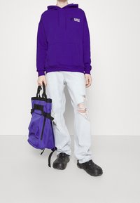 Sudadera morada con bolsillo frontal y texto impreso, combinada con jeans rasgados de color azul claro y zapatos negros sin cordones, sosteniendo una bolsa morada con correas.