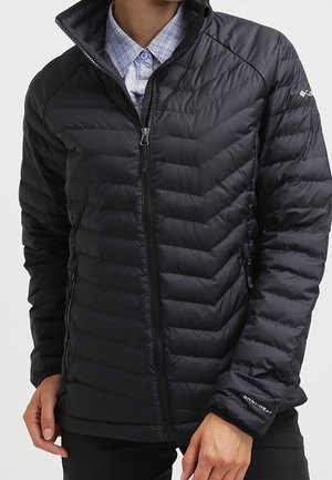 Person trägt eine schwarze gesteppte Columbia-Jacke mit Reißverschluss vorne über einem blau karierten Hemd mit Knöpfen, die Hände locker an den Seiten.