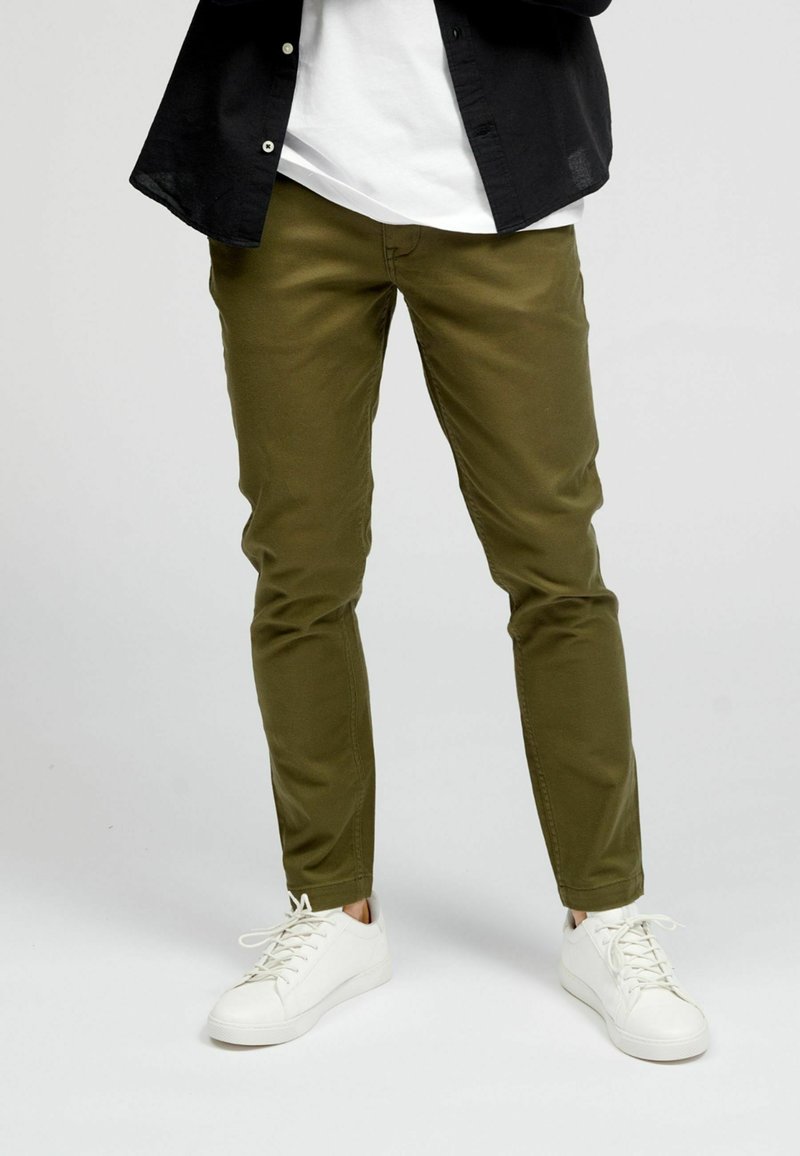 Partie inférieure du corps d'une personne portant un pantalon slim vert olive, des baskets blanches, une chemise blanche et une chemise noire ouverte à boutons.