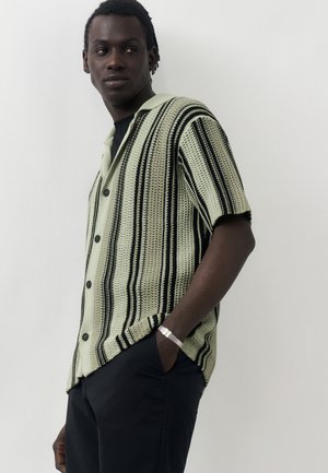 Άντρας που φοράει κοντομάνικο, πλεκτό πουκάμισο με γιακά και κάθετες ρίγες σε μαύρο και μπεζ, σκούρο παντελόνι και ασημένιο βραχιόλι, ποζάροντας πλαγίως.