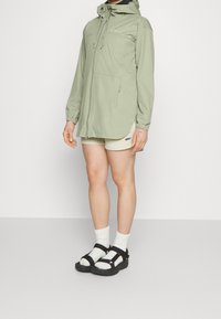 Persoon in een lichtgroene hoodie, beige shorts, witte sokken en zwarte sandaal, staand tegen een effen witte achtergrond.