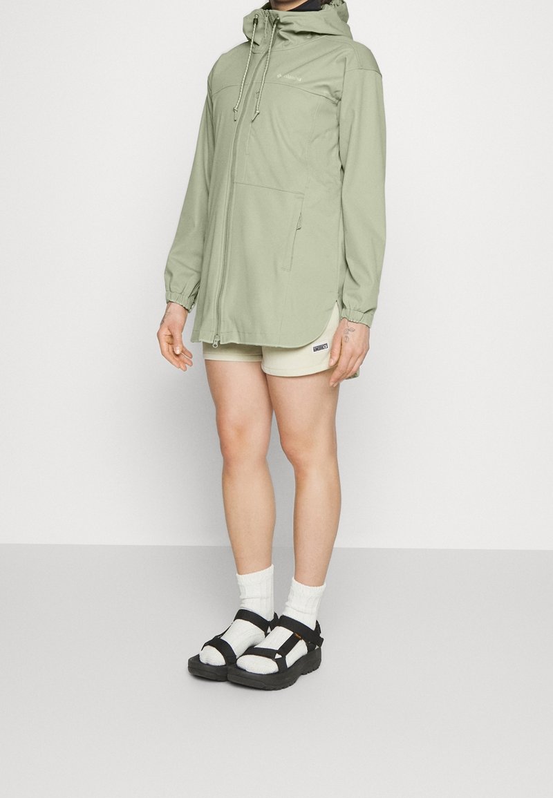 Persoon in een lichtgroene hoodie, beige shorts, witte sokken en zwarte sandaal, staand tegen een effen witte achtergrond.