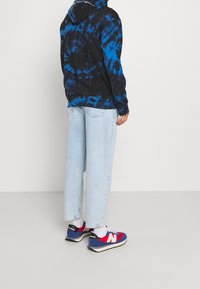 Mikina s kapucí v černo-modrém tie-dye designu, volný střih s klokánkovou kapsou. Světlé volné džíny. Boty New Balance modré a červené s bílými detaily.
