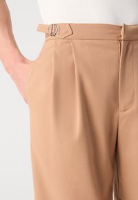 Pantalones de tiro alto en color beige con una textura suave, que presentan un pasador para cinturón y una hebilla de metal plateado. La parte delantera con pliegues añade estructura.