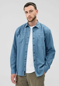 Een man met kort haar en een baard draagt een blauw overhemd over een witte T-shirt en poseert tegen een grijze achtergrond.