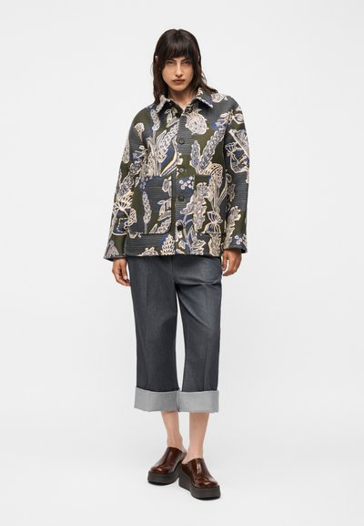 Γυναίκα που φοράει ένα σκούρο σακάκι με λουλουδάτο μοτίβο, γκρι παντελόνια με φαρδιά ποδαράκια και καφέ πλατφόρμες τύπου slip-on, στέκεται μπροστά σε έναν απλό φόντο.