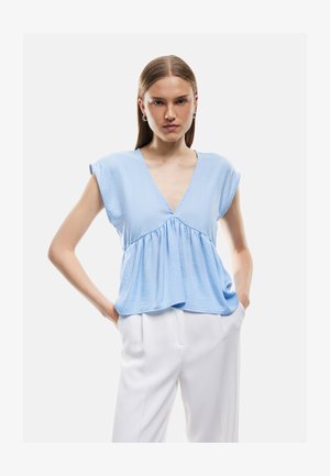 Młoda kobieta z długimi włosami, nosząca srebrne kolczyki koła, jasnoniebieską bezrękawnikową bluzkę z dekoltem w serek i białe spodnie, stojąca z rękami w kieszeniach.