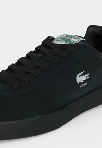 Μαύρο sneaker Lacoste με μαύρα κορδόνια, λευκό λογότυπο κροκοδείλου, κείμενο "EST. 1933" και ετικέτα γλώσσας με πράσινο και λευκό λογότυπο.