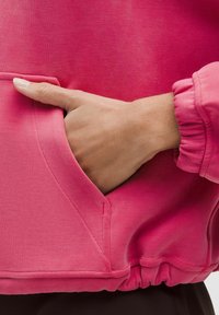 Fuksija pink majica s kapuljačom koja ima bočnu džep i elastične manžete, izrađena od glatkog materijala s mekanom teksturom i minimalističkim dizajnom.