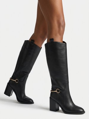 Botas negras de cuero hasta la rodilla con tacones cuadrados, detalle de metal en tono dorado con diseño de estribo de caballo, usadas sobre piernas desnudas sobre un fondo claro y sencillo.