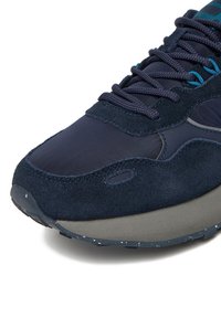 Tumši zila tenisa kurpe, kas apvieno gludu neilonu un teksturētu zamšu, ar gumijas zolīti, atbilstošām šņorēm un smalku raibu detaļojumu.