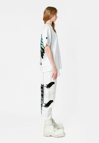 Camiseta blanca de gran tamaño con un gráfico de mariposa negra y turquesa, combinada con pantalones blancos que presentan motivos de alas negras y correas, completada con botas blancas.